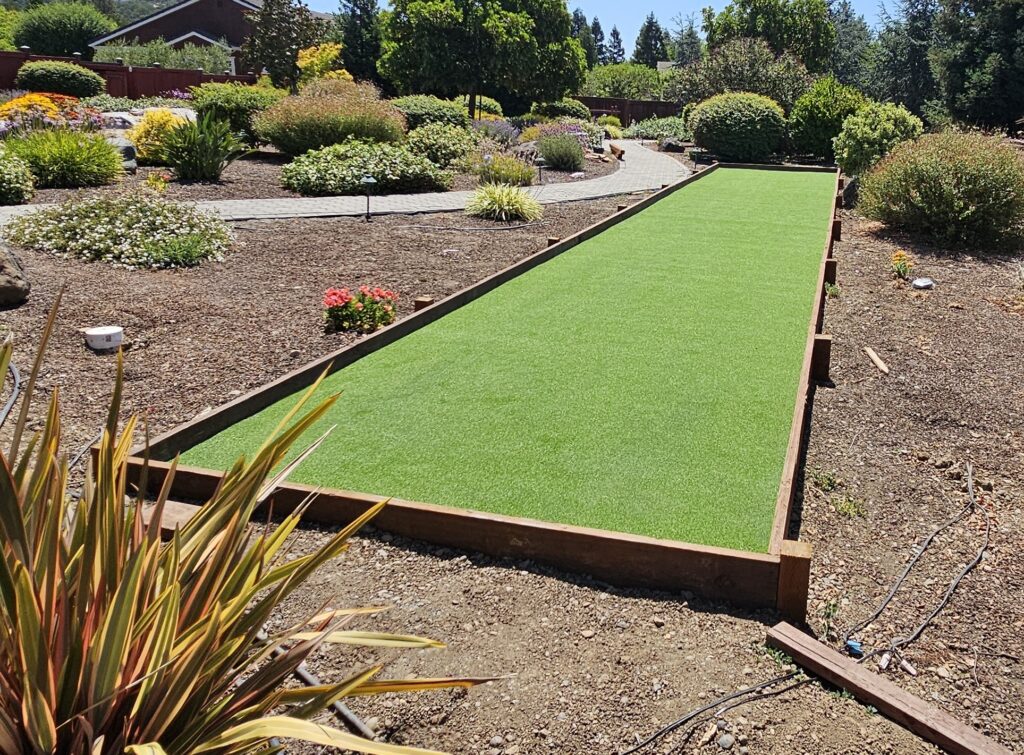 Synthetic turf bocce court with timber sideboards, bordered by drought-tolerant planting beds and a paver path in a landscaped backyard